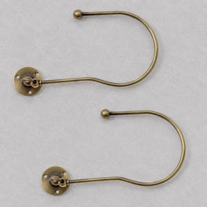 May include: Two antique brass-finished metal curtain tieback hooks. Each hook features a circular wall-mounting plate with three screw holes and a curved, J-shaped arm that terminates in a smooth, rounded ball finial at the end.
