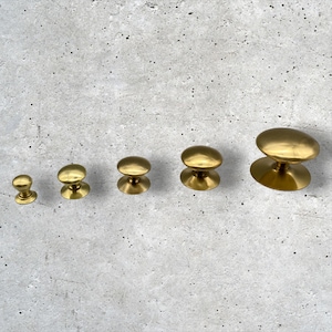 Könnte beinhalten: Fünf Messing-Schrankknöpfe in verschiedenen Größen sind in einer Reihe vor einem strukturierten grauen Hintergrund angeordnet. Die Knöpfe haben eine abgerundete Oberseite und einen kreisförmigen Sockel mit glänzendem Metall-Finish.