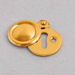 以下が含まれることがあります： 光沢のあるゴールドトーンの金属製鍵穴カバー。円形のヒンジ付きフラップが丸いベースプレートに取り付けられています。ベースプレートには中央の鍵穴と、ドアや家具に取り付けるための小さなネジ穴が1つ開いています。