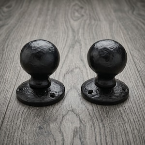 以下が含まれることがあります： 球形でテクスチャのあるハンマー仕上げが施された2つの黒いドアノブ。各ノブには取り付け用の2つのネジ穴が見える円形のベースプレートがあり、全体的に一貫した暗いマットな外観を示しています。