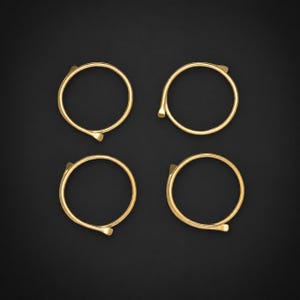 Peut inclure: Quatre anneaux circulaires dorés disposés en une grille de deux par deux. Chaque anneau présente une bande fine et lisse avec une petite saillie distincte et légèrement pointue là où les extrémités du fil métallique se rejoignent.