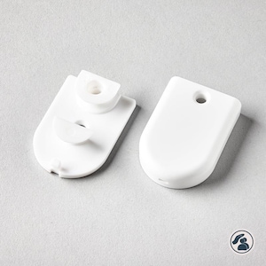 Puede incluir: Dos componentes de plástico blanco para la tensión de cordones de persianas. Una pieza presenta una base plana con una guía semicircular sobresaliente y un orificio central, mientras que la otra es una placa de cubierta lisa y redondeada.