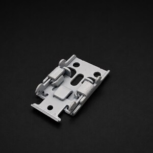 May include: A light gray metal mounting bracket featuring a rectangular base with integrated spring-loaded clips, multiple circular and elongated screw holes, and a hinged mechanism designed for secure attachment of hardware components.