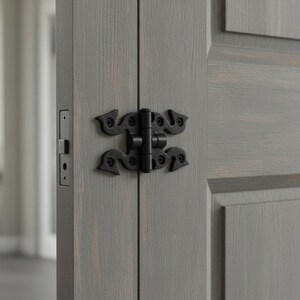May include: A grey wooden door featuring a black metal decorative hinge with a scroll-like design and a rectangular metal latch plate installed on the edge. The door has a visible wood grain texture and a panelled design with a matte finish.