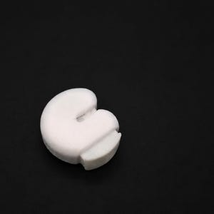 May include: A small, white, rounded object with a smooth, matte finish. The item features a distinct C-shaped or crescent-like silhouette with a flat, protruding tab on one side, creating a compact and solid form against a dark background.