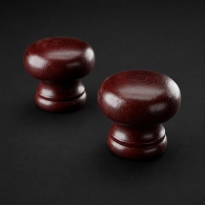 May include: Two dark reddish-brown wooden cabinet knobs with a smooth, rounded top and a tiered, circular base. These functional hardware pieces feature a polished finish that highlights the natural wood grain texture across their surfaces.