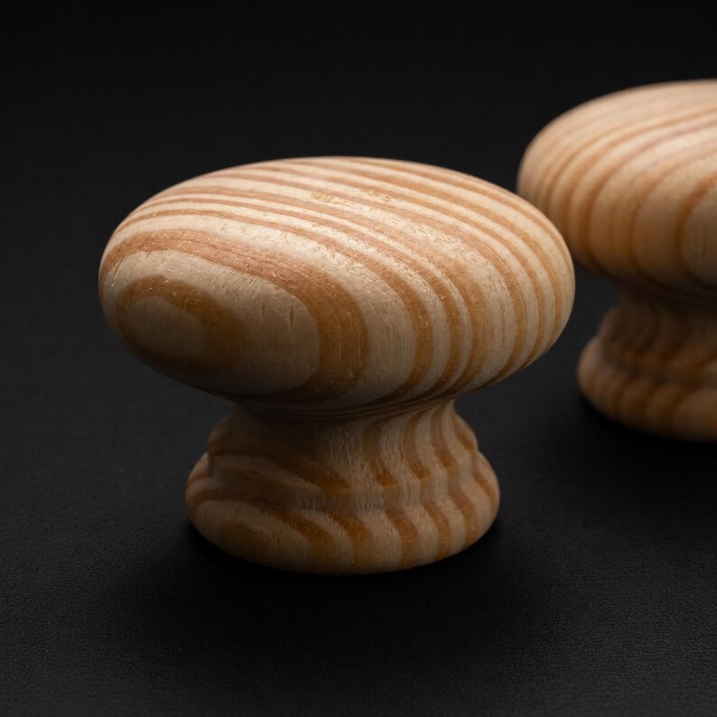 May include: Two round cabinet knobs made of light-coloured wood with a visible natural grain pattern. Each knob features a smooth, rounded top surface and a tapered base, showcasing the tan and light brown concentric rings of the wood grain.
