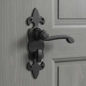 May include: A matte black door handle set featuring a decorative backplate with fleur-de-lis style ends. The assembly includes a horizontal lever handle and a small thumb-turn privacy lock mechanism, secured to the door with four visible screws.