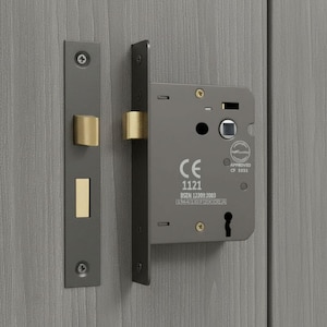 May include: A dark gray metal mortise lock set with brass-colored latch and deadbolt components. The lock body features the CE 1121 mark, BSEN 12209:2003 text, and a keyhole cutout. The strike plate is long and narrow with two visible screw heads.