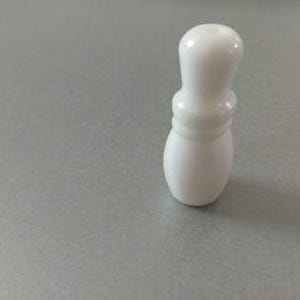 May include: A small, solid white ceramic object with a smooth, glossy finish. The item features a rounded top, a narrow neck section, and a wider, bulbous base, resembling the shape of a miniature bowling pin or a decorative game piece.