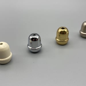 以下が含まれることがあります： 一列に並んだ4つのどんぐり型のフィニアル。1つ目はマットなオフホワイト、2つ目はポリッシュクローム、3つ目はポリッシュブラス、4つ目はブラッシュドニッケルです。各フィニアルの上部中央には小さな穴が開いています。
