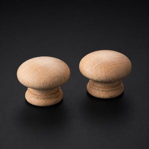 以下が含まれることがあります： 滑らかなキノコ型のトップと段差のある円形のベースを備えた、未塗装の明るい茶色の木製キャビネットノブ2個。両方のノブの表面には自然な木目がはっきりと見え、明るい茶色の色合いと質感が特徴です。