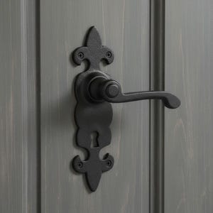 Pode incluir: Um conjunto de maçaneta de porta em preto mate com acabamento texturizado e placa decorativa com detalhes inspirados na flor-de-lis no topo e na base. O conjunto inclui uma alavanca curva e uma fechadura central, fixada com quatro parafusos.