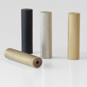 Op de afbeelding: Vier cilindervormige metalen grepen met een textuur van gekartelde ruitpatronen. Eén greep is matzwart, één is zilverkleurig en twee zijn goudkleurig. Elk onderdeel heeft een klein rond gat aan de bovenzijde voor montage.