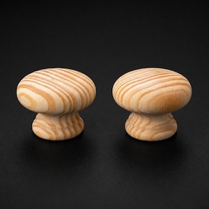 May include: Two round cabinet knobs made of light-coloured wood with a visible natural grain pattern. Each knob features a smooth, domed top surface and a slightly flared base, showcasing the organic tan and cream swirls of the wood texture.