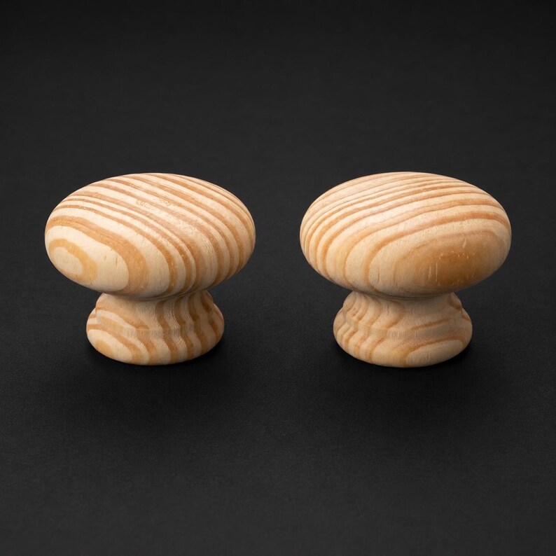 May include: Two round wooden knobs featuring a natural light tan colour with visible dark brown wood grain patterns. Each knob has a smooth, domed top surface and a slightly flared base, showcasing the organic texture and rings of the wood material.