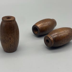 May include: Three barrel-shaped wooden beads with a dark brown finish and visible wood grain. Each bead features a central hole running through the length of the cylinder, with one bead standing upright and two beads lying horizontally on their sides.