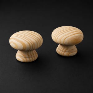 May include: Two round cabinet knobs made of light-coloured natural wood with a visible grain pattern. Each knob features a smooth, mushroom-shaped top and a tiered, circular base, showcasing the organic texture and tan tones of the wood material.