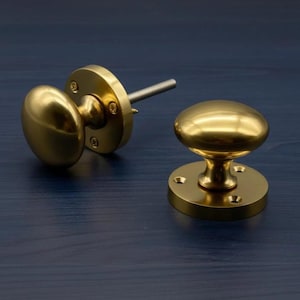 Könnte beinhalten: Zwei Messing-Schrankknöpfe mit glatter, abgerundeter Oberfläche. Jeder Knopf verfügt über eine kreisförmige Grundplatte mit drei sichtbaren Schraublöchern zur Montage. Ein Knopf ist von der Seite mit Metallstift, der andere aufrecht zu sehen.