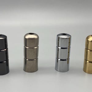 May include: Four cylindrical metal cord grips arranged in a row. Each piece features a smooth, rounded top with a central hole and two horizontal grooves along the body. The finishes include glossy black, brushed nickel, polished chrome, and brass.