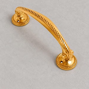 Puede incluir: Un tirador de mueble de metal dorado pulido con una barra curva de textura de cuerda. El tirador está anclado por dos placas de montaje circulares, cada una con un borde de patrón de cuerda a juego y un orificio de tornillo visible.