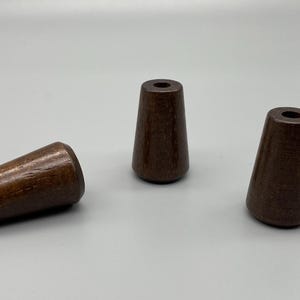 May include: Three dark brown wooden beads with a tapered, conical shape. Each bead features a smooth, polished finish and a central hole running through the length of the piece. The objects are arranged with one lying horizontally and two standing upright.