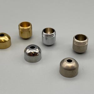 Peut inclure: Un ensemble de six petits capuchons métalliques aux finitions variées, incluant du laiton poli, du nickel brossé et du chrome. Chaque capuchon présente un sommet arrondi avec une ouverture centrale et une base cylindrique filetée.