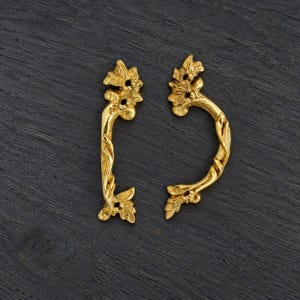 Könnte beinhalten: Zwei goldfarbene Schubladengriffe aus Metall mit einem verzierten, blattartigen Design. Jeder Griff hat eine geschwungene, längliche Form mit detaillierten botanischen Verzierungen und zwei sichtbaren Schraublöchern zur Montage an Möbeln.