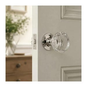 May include: A clear, faceted glass door knob with a polished chrome base and circular mounting plate, installed on a light-colored paneled door. The hardware includes a rectangular metal latch plate visible on the door edge.
