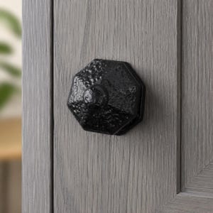 Op de afbeelding: Een zwarte achthoekige kastknop met een getextureerde, gehamerde oppervlakteafwerking. De knop is gemonteerd op een lichtgrijze houten kastdeur met een zichtbaar verticaal houtnerfpatroon, wat zorgt voor een contrast met het donkere beslag.