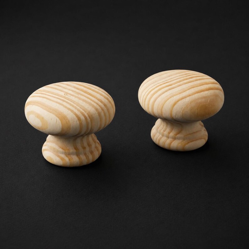 May include: Two round cabinet knobs made of light-coloured natural wood. Each knob features a smooth, mushroom-shaped top with visible wood grain patterns in shades of tan and cream, supported by a smaller, tiered circular base for mounting.