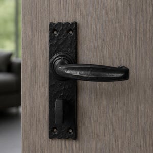 May include: A black door handle set with a hammered texture on a rectangular backplate. The hardware features a horizontal lever handle and a thumb turn lock mechanism, mounted on a light brown wood grain door with visible vertical texture.