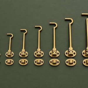 May include: A set of seven brass cabin hooks arranged in increasing order of size. Each hook features a hinged mounting plate and a separate eyelet plate, both with two screw holes. The metal has a polished gold-tone finish and a curved hook end.