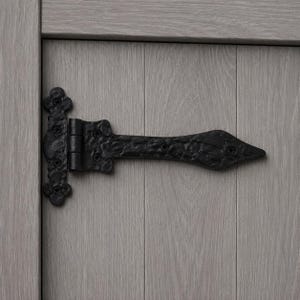 May include: A black metal T-strap hinge with a textured, hammered finish and a pointed end, secured with visible screws to a light grey, vertical-planked wooden surface. The hinge features a decorative, ornate base plate with four screw holes.