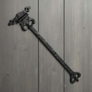 Könnte beinhalten: Ein schwarzes Metall-Langband mit texturierter Oberfläche. Das Scharnier verfügt über eine dekorative, pfeilförmige Montageplatte mit vier Schraublöchern und einen langen, geraden Arm, der in einer stilisierten Lilien-Endspitze endet.