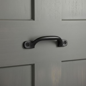 May include: A matte black metal drawer pull handle with a curved arch shape, secured by two visible screws. The handle is mounted horizontally onto a flat, gray-painted wooden surface featuring a visible vertical and horizontal panel seam.