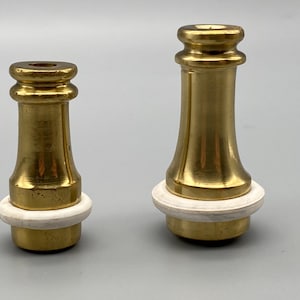 Può includere: Due portacandele in ottone di diverse altezze, ciascuno caratterizzato da una finitura metallica lucida e da una base ad anello circolare bianca e liscia. Il design presenta una sezione superiore svasata e un corpo cilindrico riflettente.