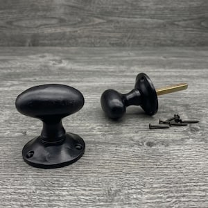 May include: Two matte black oval door knobs with circular mounting plates. One knob is shown from the front, while the other is shown from the side with a brass-colored spindle extending from the back. Four small black screws are included.