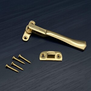 May include: A polished brass window casement fastener with a matching strike plate and four mounting screws. The hardware features a smooth, metallic finish with a curved handle design and pre-drilled holes for installation on window frames.