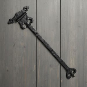 May include: A black metal strap hinge with a textured finish, featuring a decorative fleur-de-lis style end and a matching hinge plate with pre-drilled screw holes. The long, narrow arm extends from the ornate hinge mechanism, providing a functional and detailed hardware component for doors or gates.
