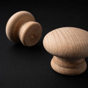 以下が含まれることがあります： 滑らかで丸みを帯びたトップと段差のある円筒形のベースを持つ、未塗装の明るい茶色の木製キャビネットノブ2個。両方のノブの表面には自然な木目がはっきりと見え、明るいベージュ色とマットな質感が特徴です。