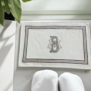 May include: White rectangular bath mat with a decorative border in black and gold. The mat features an embroidered monogram in gray and gold. Two white, fluffy bath towels are in the foreground.