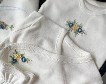 May include: A set of baby clothes, including a onesie, bib, and mittens, in a soft white fabric. Each item features delicate embroidery of blue, yellow, and green floral designs. The set is designed for newborns and infants.
