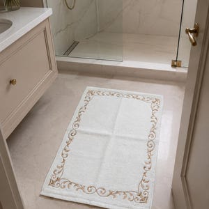 May include: A rectangular white bath mat featuring a decorative gold-toned embroidered border with a repeating scrollwork pattern. The mat has a soft, textured surface and is positioned on a light-colored tiled floor in a bathroom setting.