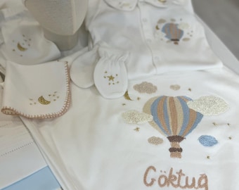 May include: A white baby clothing set featuring embroidered designs of clouds, stars, and a hot air balloon in blue and brown. The set includes a hat, blanket, shirt, bib, and mittens. The blanket has the name "Göktuğ" embroidered on it.