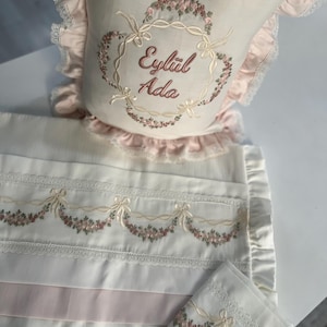 Puede incluir: Juego de ropa de cama para bebé en blanco y rosa pálido, con guirnaldas florales bordadas y ribetes de encaje. El juego incluye una almohada con el nombre "Eylül Ada" bordado en una corona floral, una manta y una funda de almohada pequeña.