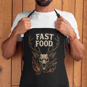 Peut inclure: Tablier noir avec un motif de crâne de cerf, flèche et flammes. Le texte "FAST FOOD" est affiché au-dessus du crâne. Le tablier a une grande poche avant et des bretelles noires.