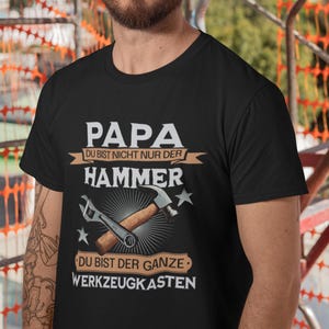 Könnte beinhalten: Ein schwarzes T-Shirt mit dem Text "PAPA DU BIST NICHT NUR DER HAMMER DU BIST DER GANZE WERKZEUGKASTEN" und Werkzeugillustrationen. Die Person im Bild trägt einen weißen Bauhelm.