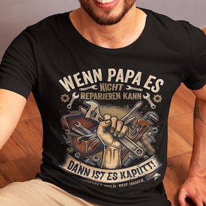 May include: Black t-shirt with a graphic design featuring tools and the German text "Wenn Papa es nicht reparieren kann, dann ist es kaputt!" A hand holds wrenches and other tools. The shirt is worn by a person.