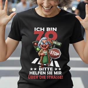 Könnte beinhalten: Schwarzes T-Shirt mit der Aufschrift "ICH BIN 70" und einer Karikatur einer älteren Frau, die einen Stock und ein Stoppschild hält. Das Shirt hat auch den Text "BITTE HELFEN SIE MIR ÜBER DIE STRASSE!"